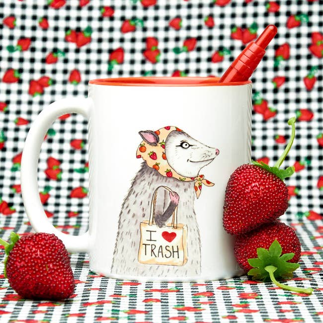 white mug with red inside and an illustration of an opossum in a scarf carrying a tote bag that says 