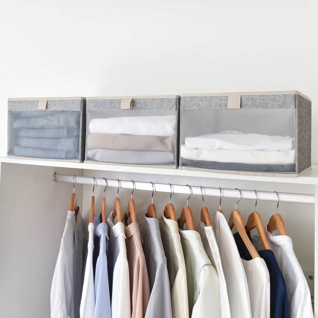 Three fabric storage bins filled with neatly folded clothes sit above a closet rail with hanging shirts