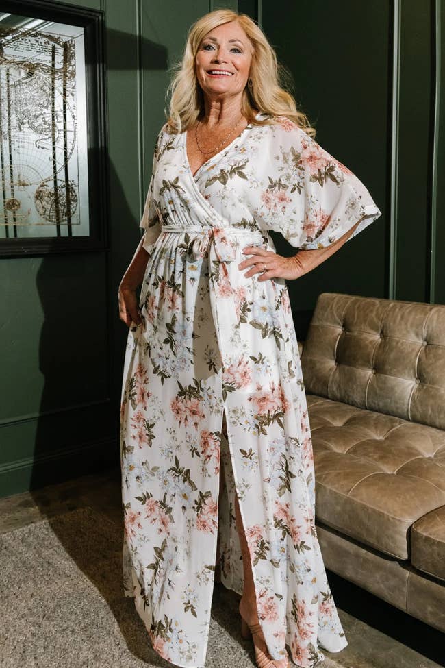 Model wearing maxi white and pink floral maxi dress with bottom slit, v-neckline, flowy sleeves, and adjustable waist tie standing in front of brown leather couch