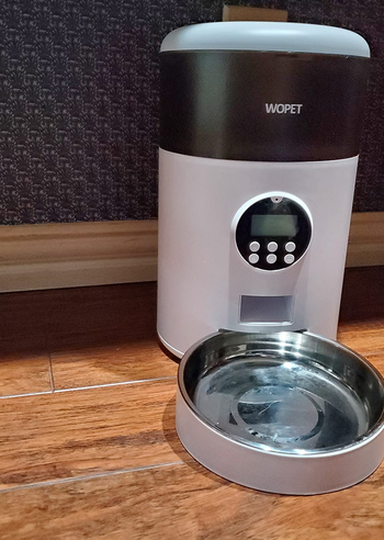 Reviewer image of black and white automatic cat feeder with personalization screen and white and silver bowl under dispenser on hardwood floor