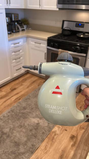 Hand holds a Bissell SteamShot Deluxe cleaner successful  a kitchen. The instrumentality  is pointed distant  from the camera, targeting household   cleaning tasks