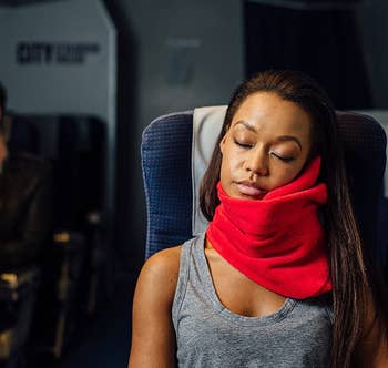 model wears bright red wrap-around Trtl neck pillow while sleeping in plane seat