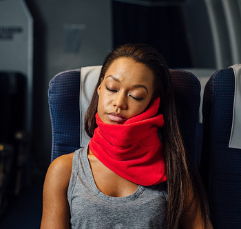 model wears bright red wrap-around Trtl neck pillow while sleeping in plane seat
