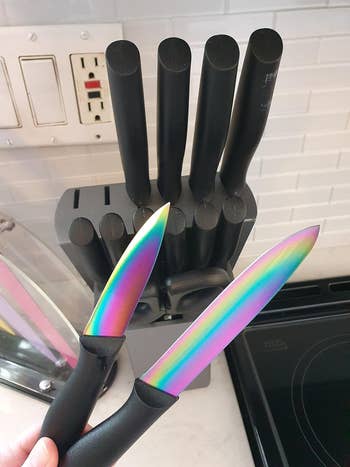 A hand holding two knives with iridescent blades from a knife block in a kitchen setup, suggesting product highlight for shopping