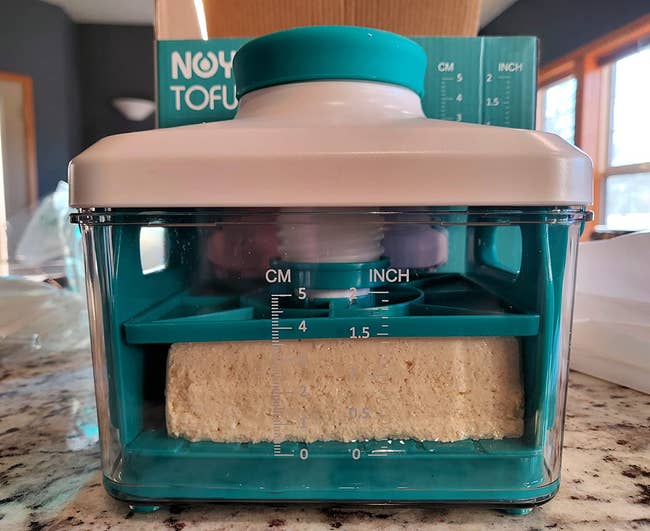 reviewer photo of the teal tofu press with measurement markings on its side pressing the liquid out of a block of tofu
