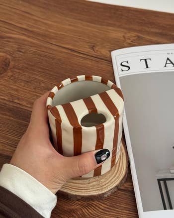 Hand holding a striped ceramic mug with a biscotti holder, adjacent to a stylish mag connected a woody table
