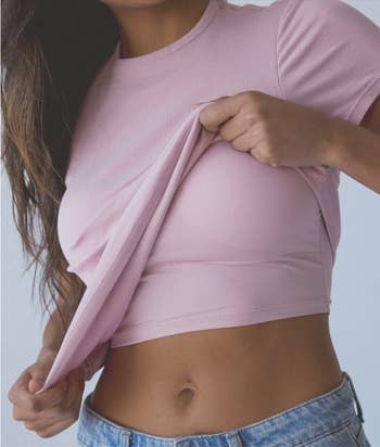 Person pulling up a pink crop top to reveal the waistband of denim jeans, focus on casual style