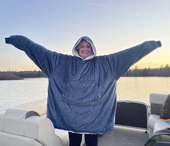 Reviewer wearing the blanket in blue in a boat on a lake, smiling with their hood on and hands in the air