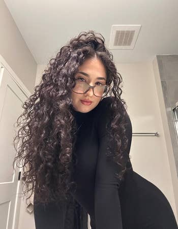 Person with agelong  curly hairsbreadth  and glasses wearing a form-fitting achromatic  outfit, posing indoors with a neutral expression