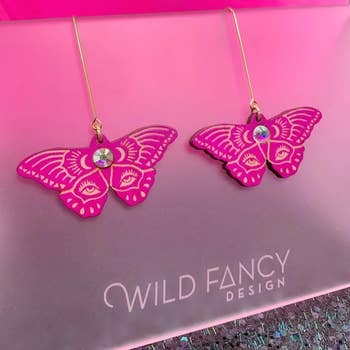 Butterfly-shaped earrings from Wild Fancy Design, featuring intricate patterns and gem accents, displayed connected a pinkish background