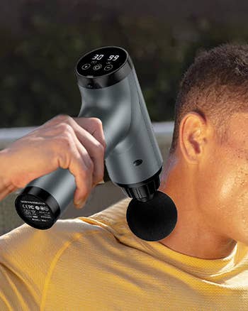 Person using a handheld massage gun on their neck, showcasing its ergonomic design and digital controls