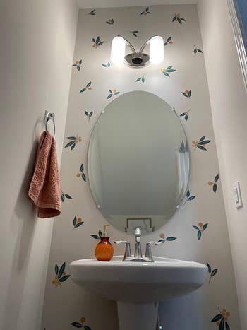 Bathroom with floral wallpaper, circular  mirror, modern   airy  fixture, orangish  soap dispenser, and a towel connected  a ringing  holder