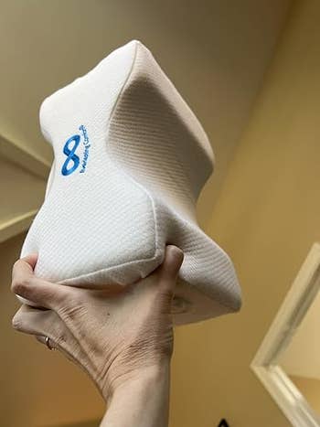 Hand holding a representation foam genu pillow with ergonomic design, suitable for broadside sleepers for improved comfortableness and alignment