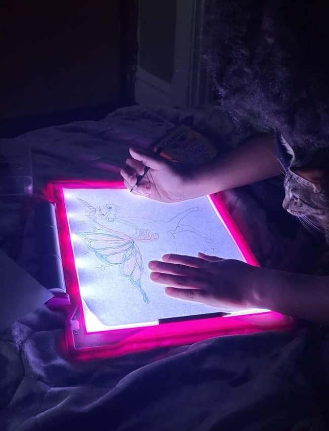 Person tracing a butterfly and flower drawing on a light-up tracing pad in a dimly lit room