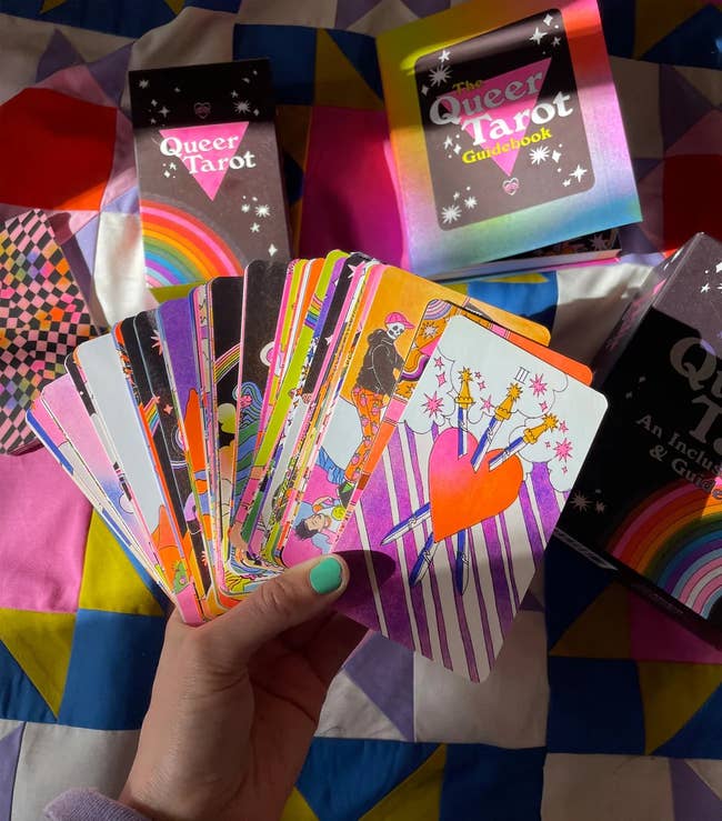 A manus  holds vibrant Queer Tarot cards with boxes successful  the background, showing playful, inclusive designs