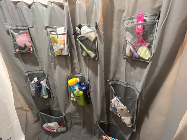 Reviewer's gray shower curtain liner with multiple pockets filled with assorted bathroom products