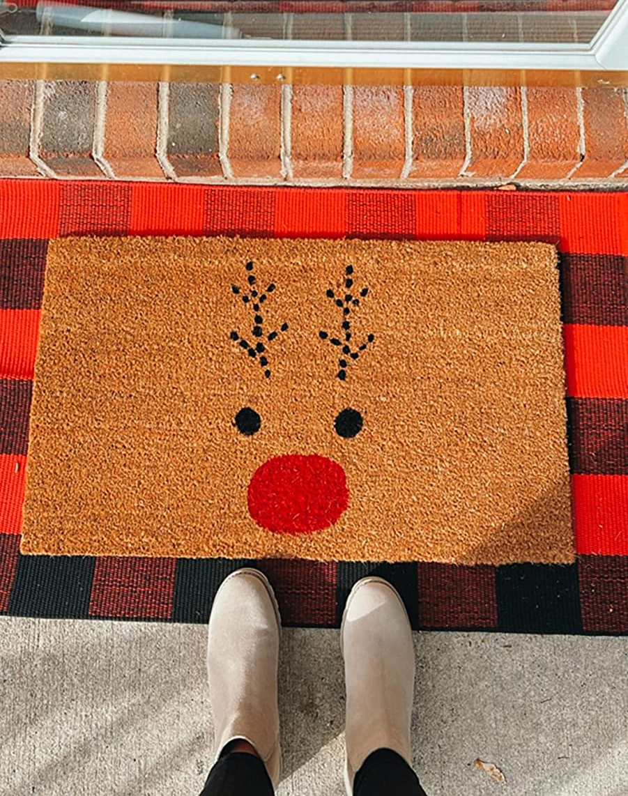 reviewer standing in front of the rudolph doormat, which is on top of a red and black buffalo plaid mat