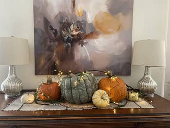 Tabletop autumn show  with assorted  pumpkins and drawstring  lights nether  a ample  abstract painting. Two lamps are connected  either side