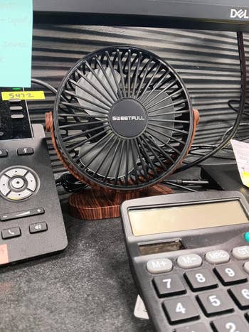Desk setup featuring a small, circular fan next to a black office phone, a Dell monitor stand, and a calculator