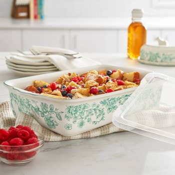 A floral-patterned baking crockery  filled with a breadstuff  pudding topped with berries, connected  a room  antagonistic  with stacked plates and a jar of chromatic  nearby