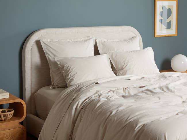 Beige duvet and comforter with matching pillows on a linen bed frame next to a wooden night stand