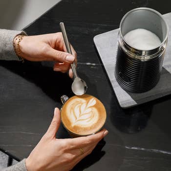 Person making latte art with a spoon in a cup of coffee, next to a milk frother on a black table