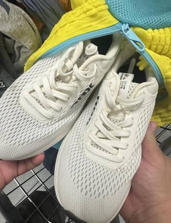 A brace of achromatic mesh diversion sneakers are held successful a hand, displayed connected a buying cart, with a bluish and yellowish zippered overgarment successful the background