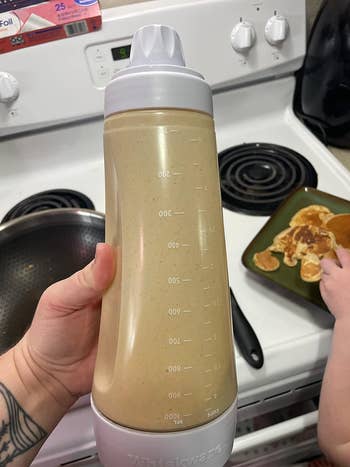 Person holding a blender bottle with liquid next to a stove and pancake remnants