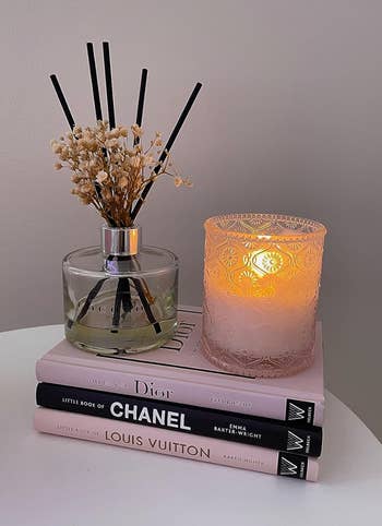 Diffuser on books by Dior, Chanel, Louis Vuitton, beside a lit candle, on a table for a cozy ambiance
