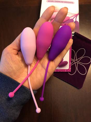 Hand holding three Kegel exercise weights next to their packaging and a storage pouch