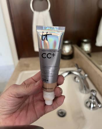 Person holding a conduit  of IT Cosmetics CC+ cream, SPF 50+, adjacent   a bath  sink