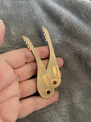 Hand holding two brass-colored, serrated-edged pocket hooks for shopping bag handles, resting on a soft fabric surface