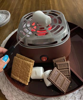 reviewer using s'mores maker to roast a marshmallow with graham crackers and chocolate in the trays