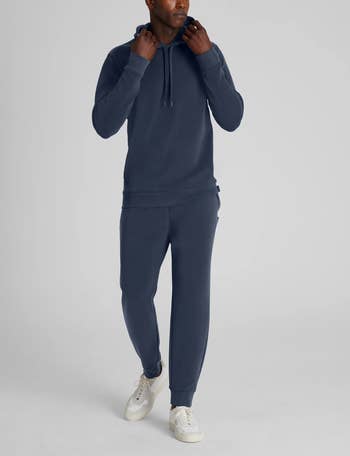 Person wearing a casual hoodie and joggers set with white sneakers, posing against a plain backdrop