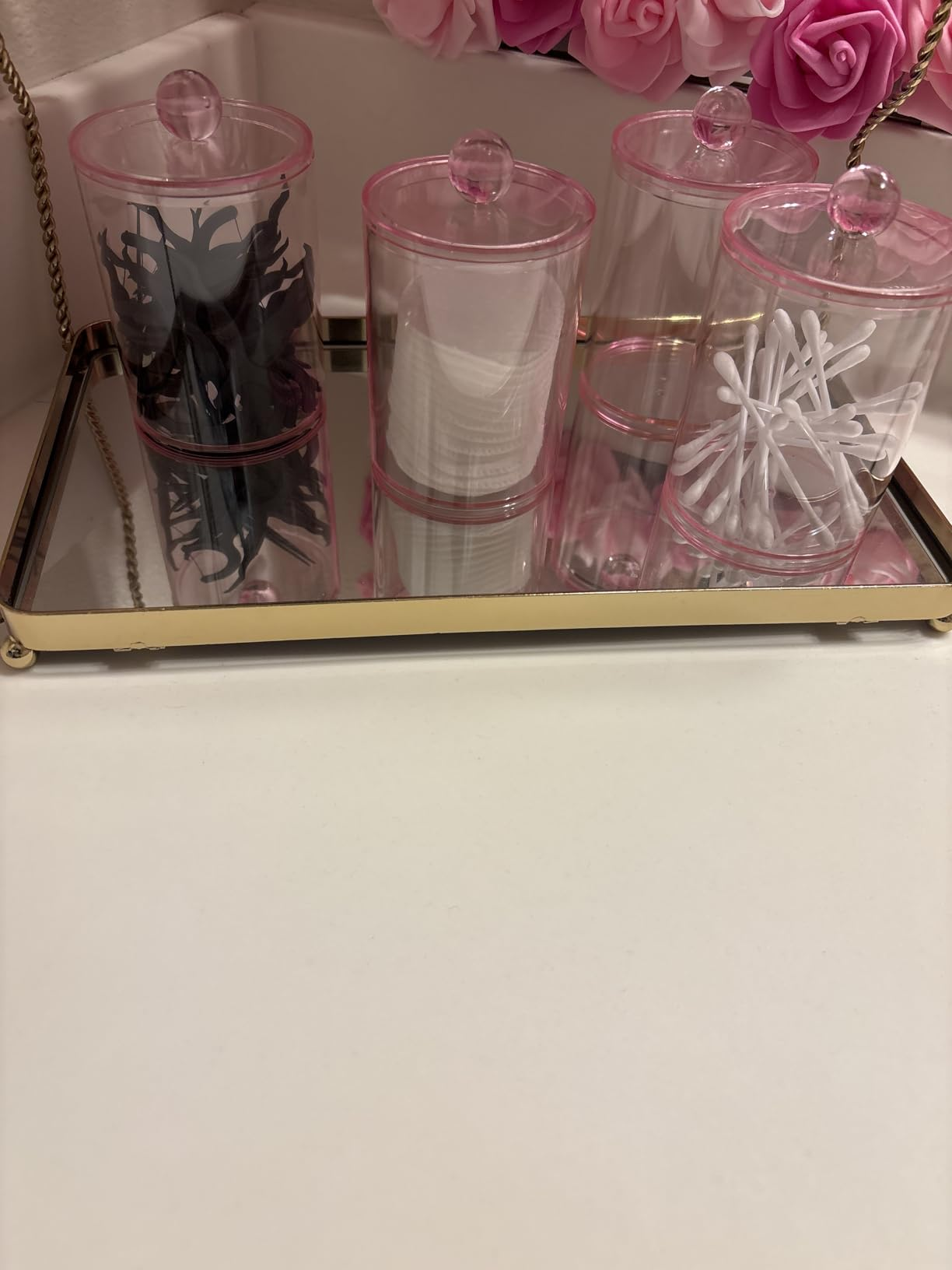 Vanity tray with clear jars holding makeup pads, cotton swabs, and hair clips, set against a background of pink roses