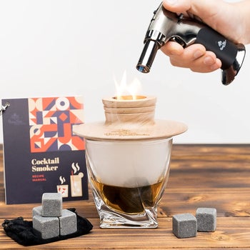 The kit with its contents laid out showing a model using a torch on a cocktail with a wooden lid 
