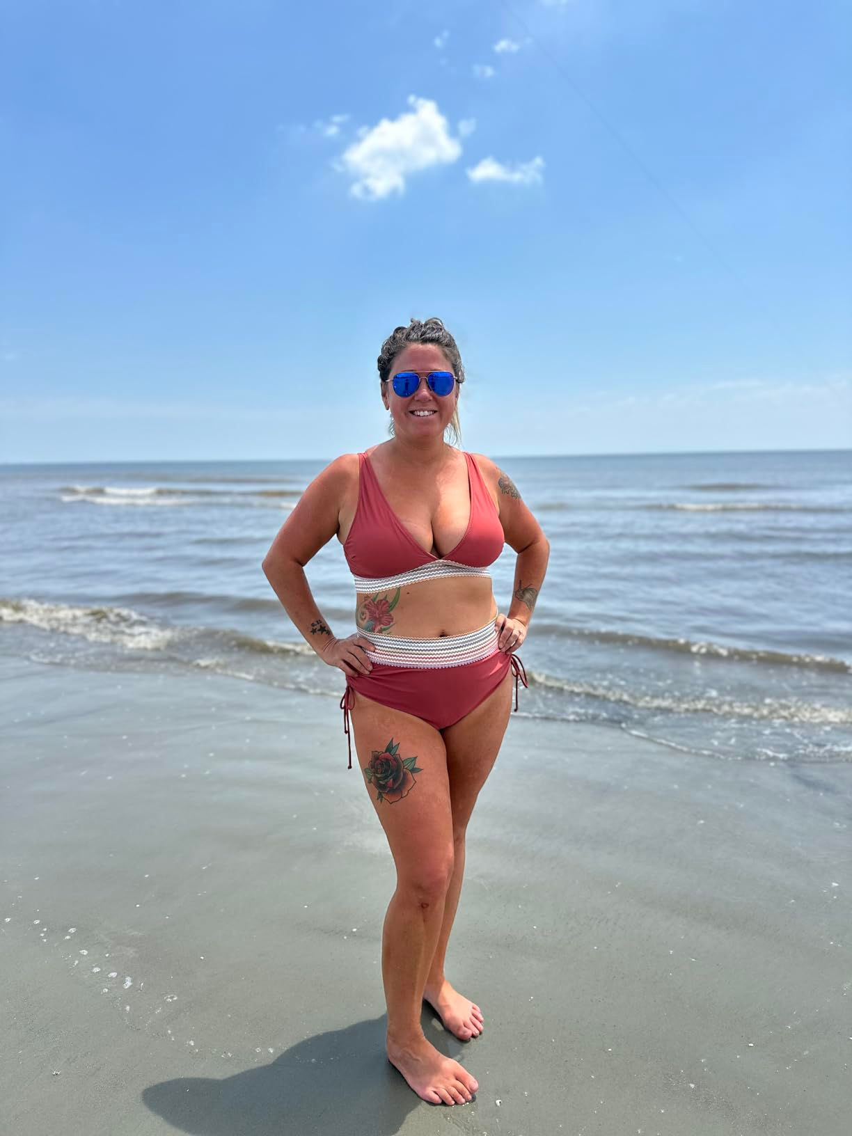 Person stands on a beach wearing a two-piece swimsuit with body tattoos visible, looking confident and smiling under a clear sky