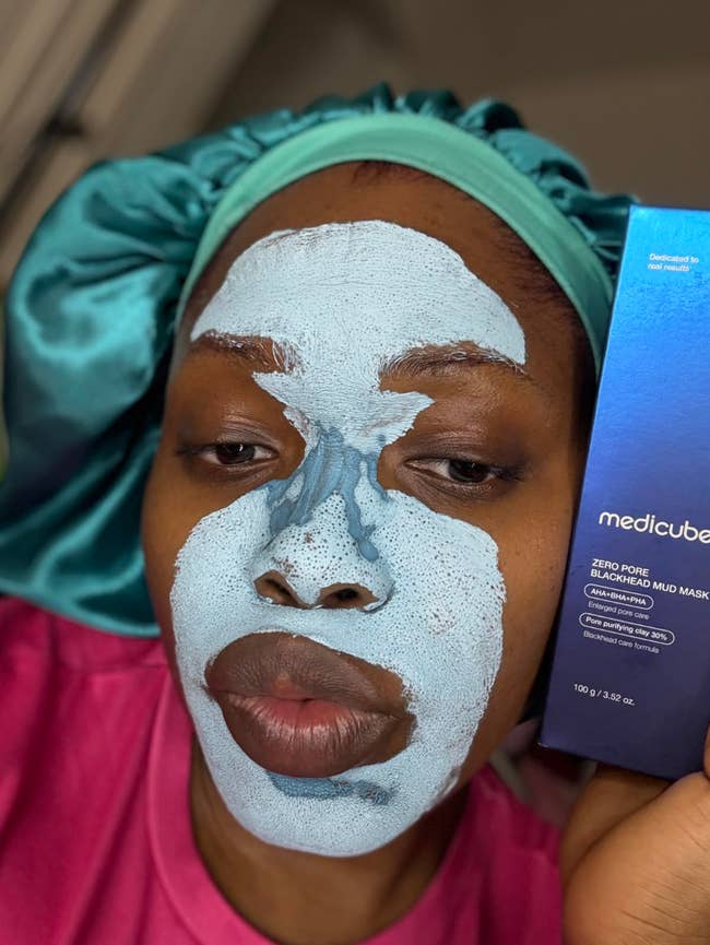 Person with a bluish look disguise holds a container of Mediecube Zero Pore Blackhead Mud Mask, wearing a silky bonnet