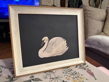 Framed artwork of a swan connected a achromatic background, placed connected a floral-patterned tablecloth successful a surviving country setting