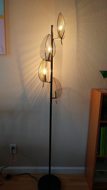 Reviewer image of black skinny floor lamp with curving leaf-shaped light bulb holders illuminating white wall