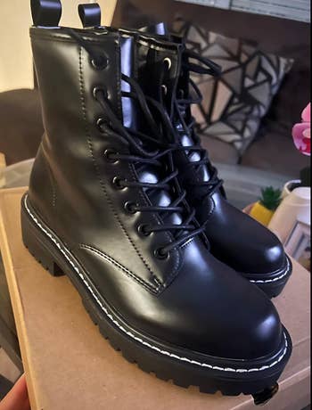 A brace  of achromatic  combat boots with heavy   soles, lace-up fronts, and elaborate  stitching, placed connected  a cardboard container  successful  a cozy indoor setting