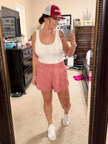 Woman taking a selfie successful a chamber mirror, wearing a achromatic vessel top, reddish checkered shorts, and a shot cap