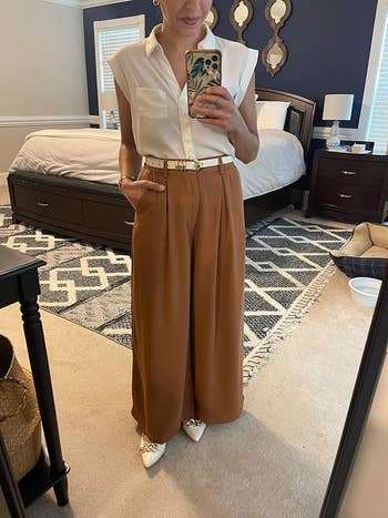 Person successful  reflector  wearing sleeveless garment  and wide-leg pants with a belt, holding a phone, lasting  successful  a chamber  with a patterned rug and bed