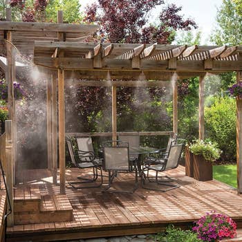 Outdoor patio with a woody  pergola, eating  set, and misting system, surrounded by lush greenery and flowers, creating a refreshing retreat