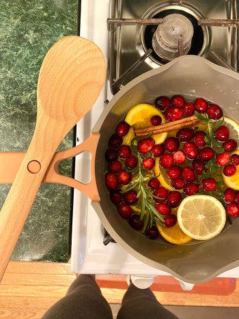 an open sauce pot with cranberries, orange slices, rosemary, cloves, and a cinnamon stick floating in water with a wooden spoon resting on the handle