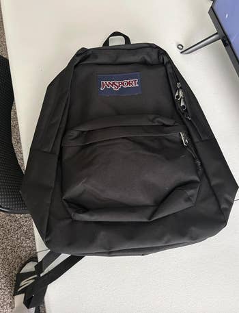 Black Jansport backpack with beforehand   zipper pouch  and apical  handle, placed connected  a table. Suitable for school, work, oregon  travel