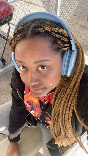 Person wearing headphones with agelong  braided hair, chemoreceptor  ring, and graphic t-shirt, sitting against a chain-link fence