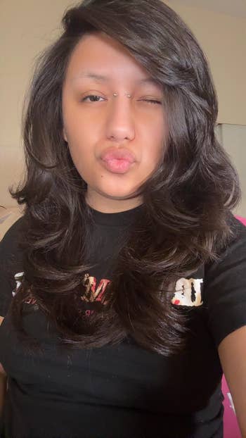Person with wavy layered hairsbreadth  poses with a wink and pout, wearing a graphic tee