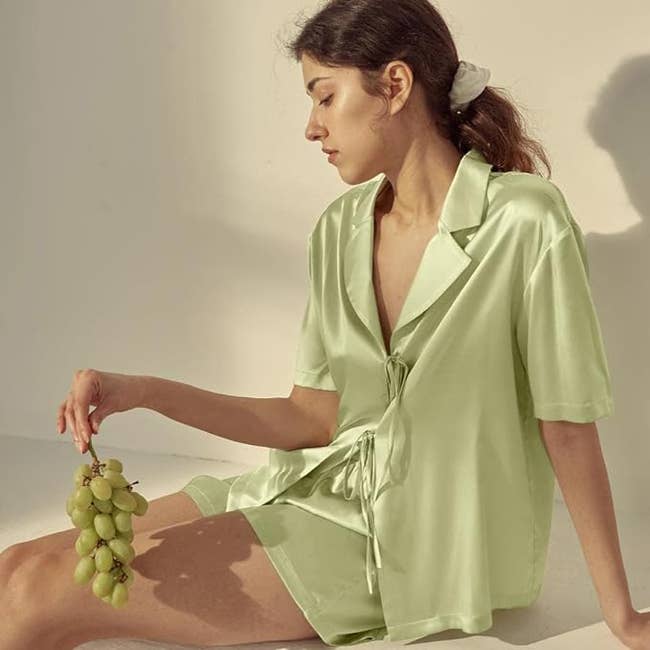 Woman in a satin robe holding grapes, seated in a relaxed pose, in a minimalist setting for a fashion retail concept