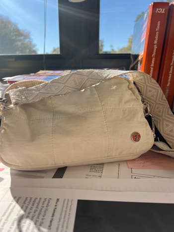 A stylish handbag with a geometric patterned strap is placed on a desk with magazines and papers, surrounded by books, suggesting a trendy design